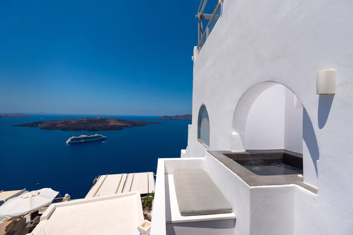A white building with a balcony overlooking the sea, a ship in the distance, and a clear blue sky.