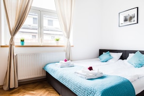Ein ordentlich bezogenes Bett mit blauer Bettwäsche, ein Fenster mit Vorhängen, ein Blumentopf und ein gerahmtes Bild an der Wand.