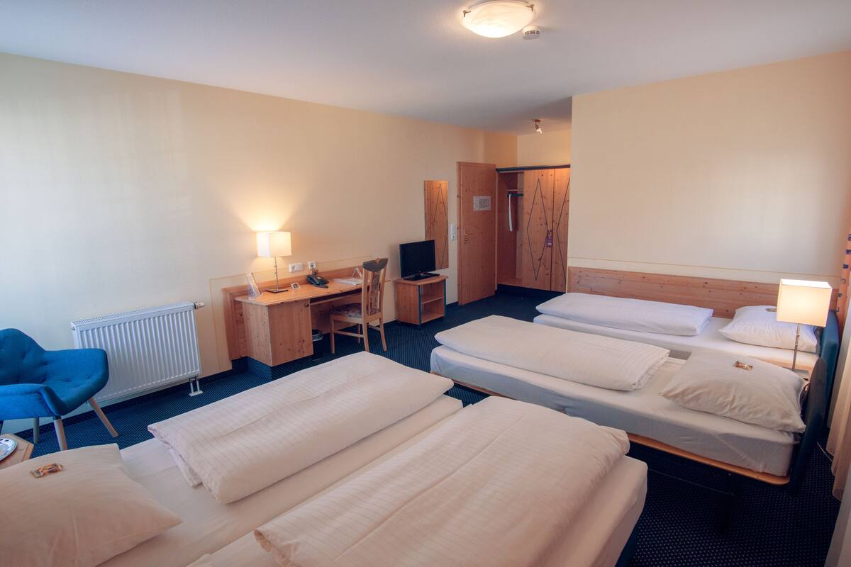 A hotel room with three beds, a desk with a computer, a chair, and a radiator.