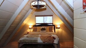 A cozy attic bedroom with a sloped ceiling, a bed with a patterned bedspread, two bedside lamps, and a window with a view of trees.