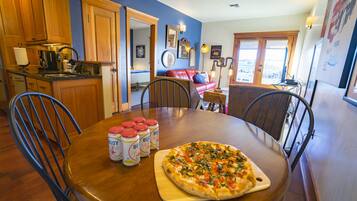 A dining area with a wooden table, chairs, and a pizza on a wooden board. There are six bottles of sauce on the table.