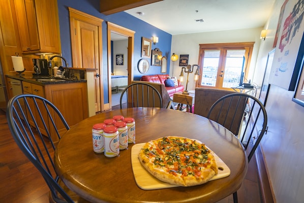 A dining area with a wooden table, chairs, and a pizza on a wooden board. There are six bottles of sauce on the table.