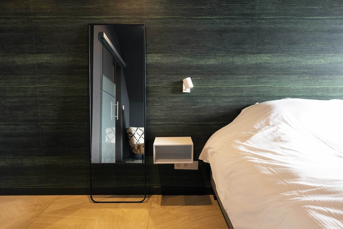 Une chambre à coucher moderne avec un grand miroir, une table de chevet avec une lampe et un lit soigneusement fait.