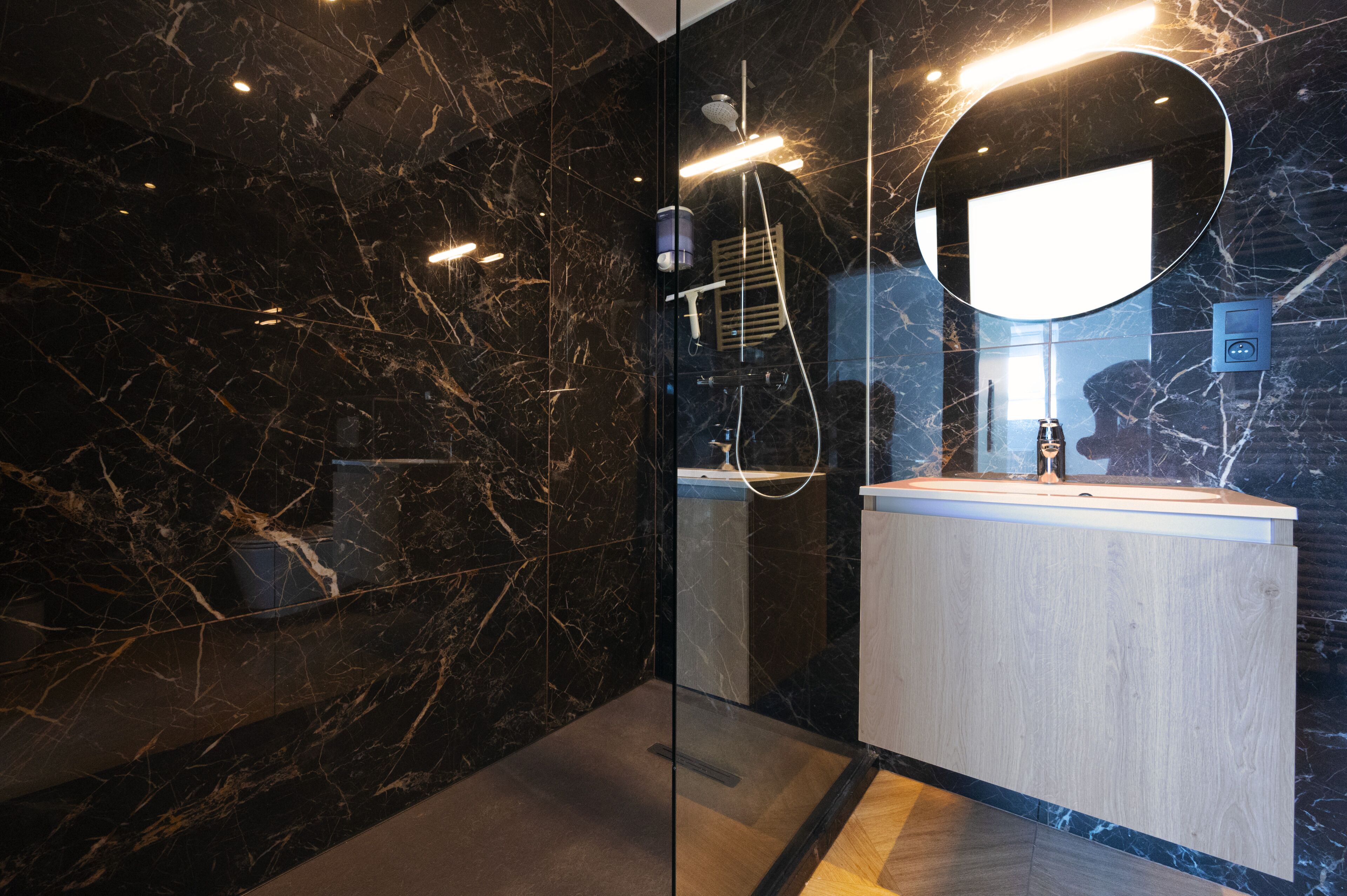 A modern bathroom with a marble wall, a wooden vanity, and a glass shower enclosure.