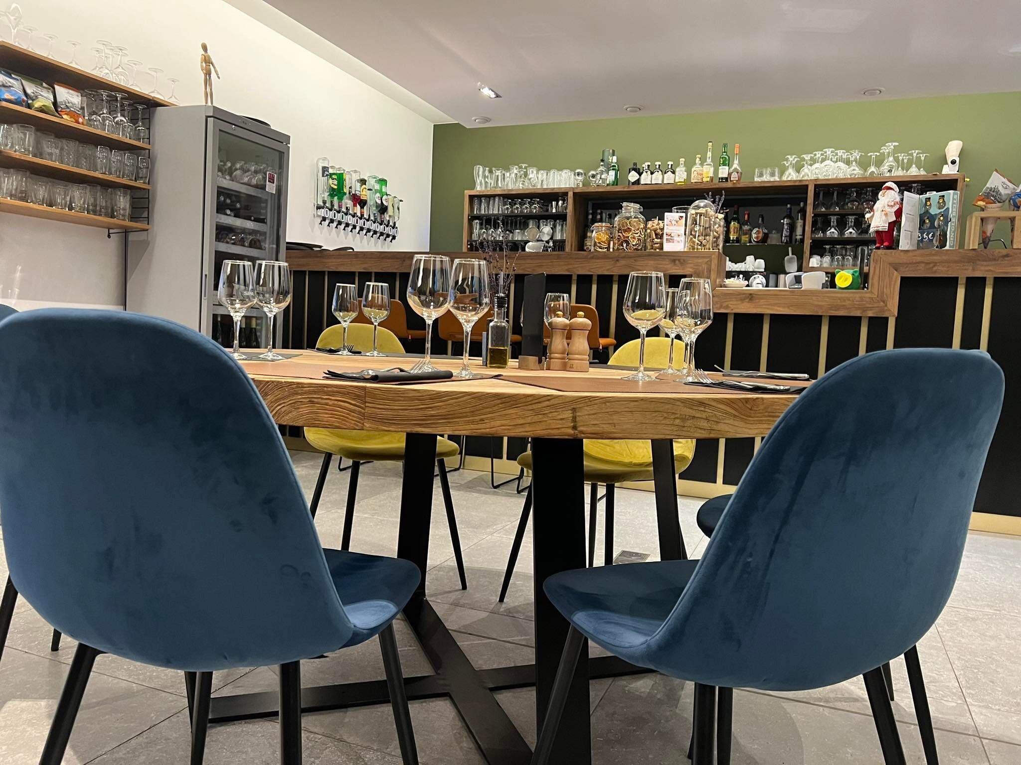 Ein Restaurantinnenraum mit Holztisch, gedecktem Esstisch, blauen Stühlen und Weinregalen mit Weingläsern und Flaschen.