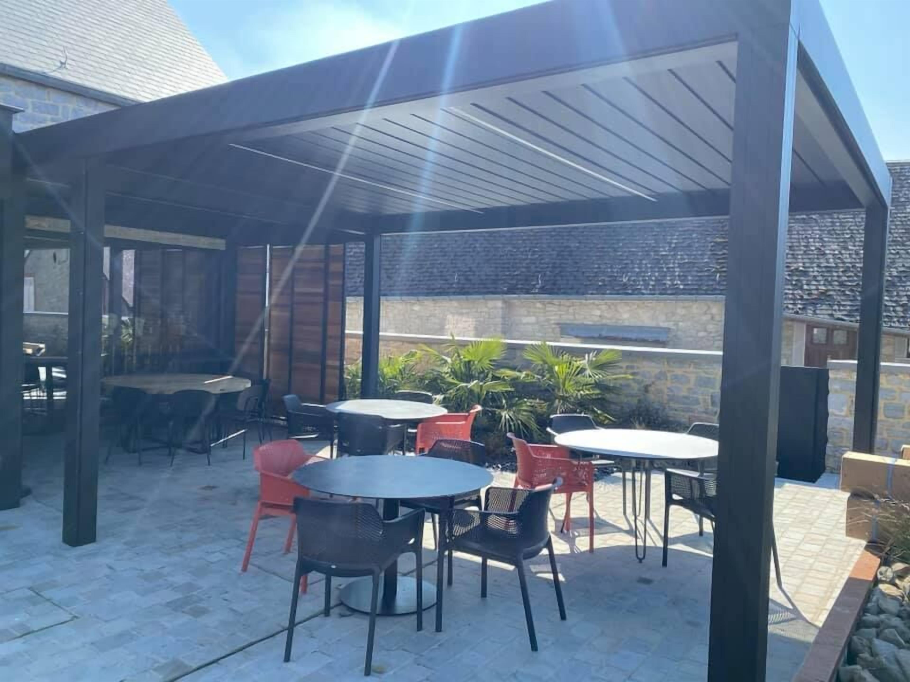 An outdoor patio with multiple tables and chairs, a covered area with a roof, and a stone wall in the background.