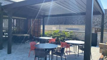 Un patio al aire libre con varias mesas y sillas, una zona cubierta con techo y un muro de piedra al fondo.