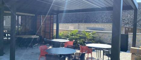 An outdoor patio with multiple tables and chairs, a covered area with a roof, and a stone wall in the background.