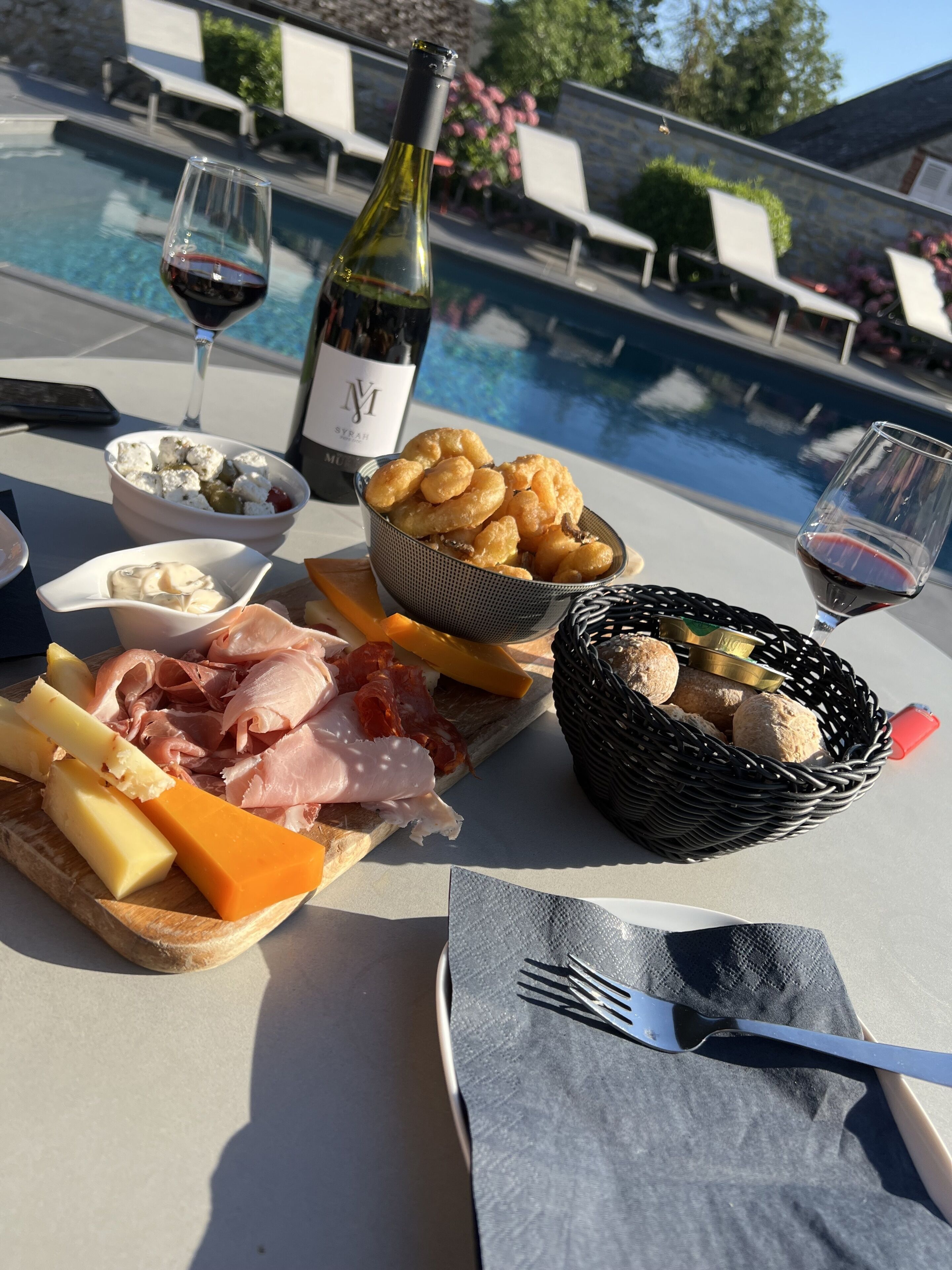 Une table dressée au bord de la piscine, sur laquelle on trouve une variété d’aliments, notamment du fromage, de la charcuterie et du pain, ainsi que des verres à vin et une bouteille.