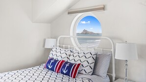 Un dormitorio con cama, almohadas, una mesita de noche, lámpara y ventana con vista al mar.