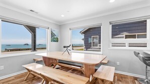 Amplia habitación con una gran mesa de madera y bancos, ventanas grandes que ofrecen vistas al océano y un diseño moderno del techo.