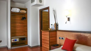 Habitación de hotel con un armario de madera, un espejo, una mesita de noche con una lámpara y un cojín rojo.