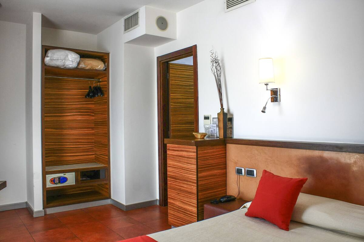 A hotel room with a wooden wardrobe, a mirror, a bedside table with a lamp, and a red pillow.