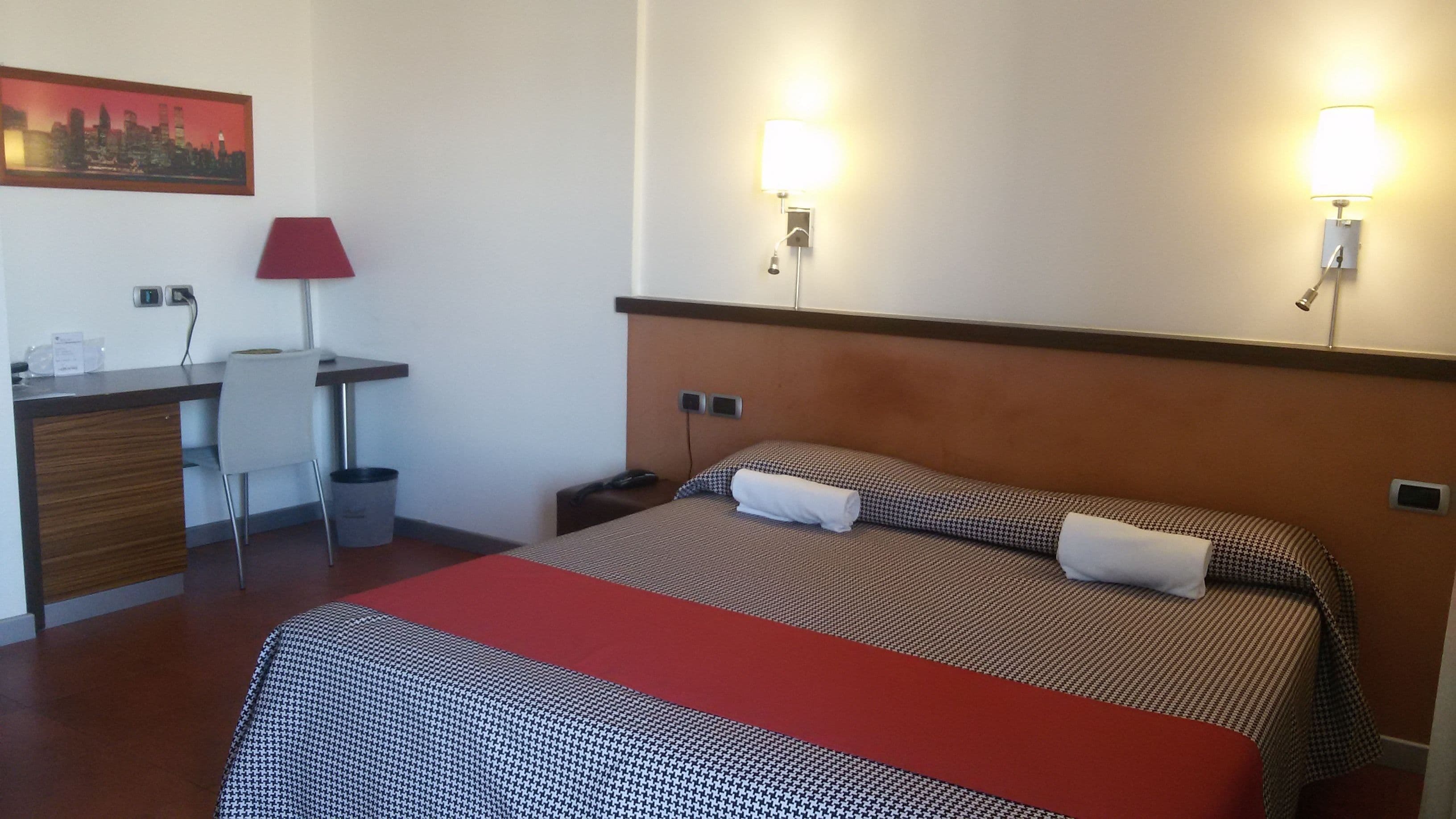 Habitación de hotel con cama, escritorio, silla, cesto de basura y dos luces fijadas a la pared.