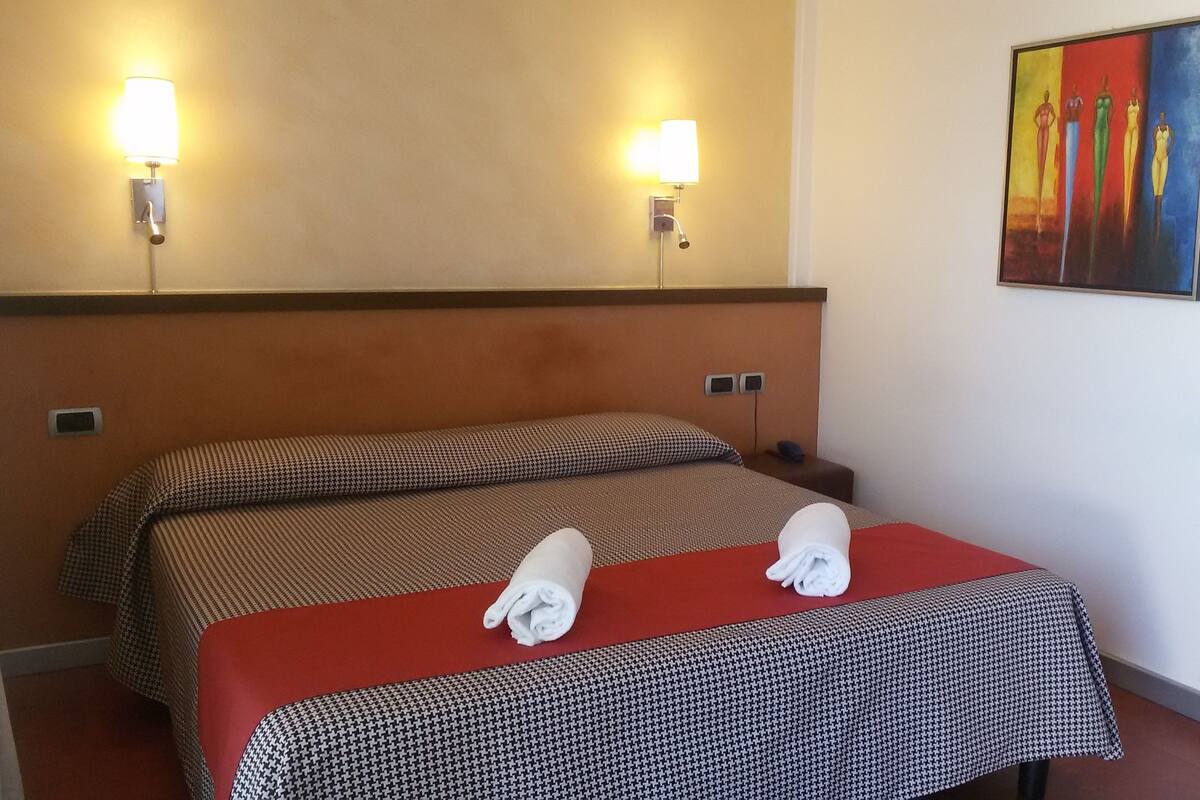 A hotel room with two beds, each with a patterned bedspread and a red blanket. There are wall-mounted lamps and a painting on the wall.