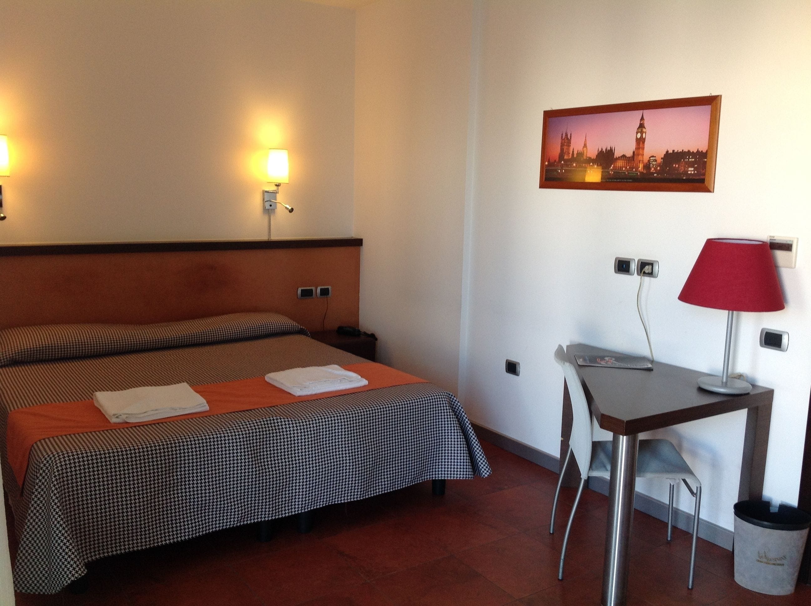 Ein Hotelzimmer mit zwei Betten, einem Schreibtisch, einem Stuhl, einer Lampe, einem Papierkorb und einem Stadtbild an der Wand.