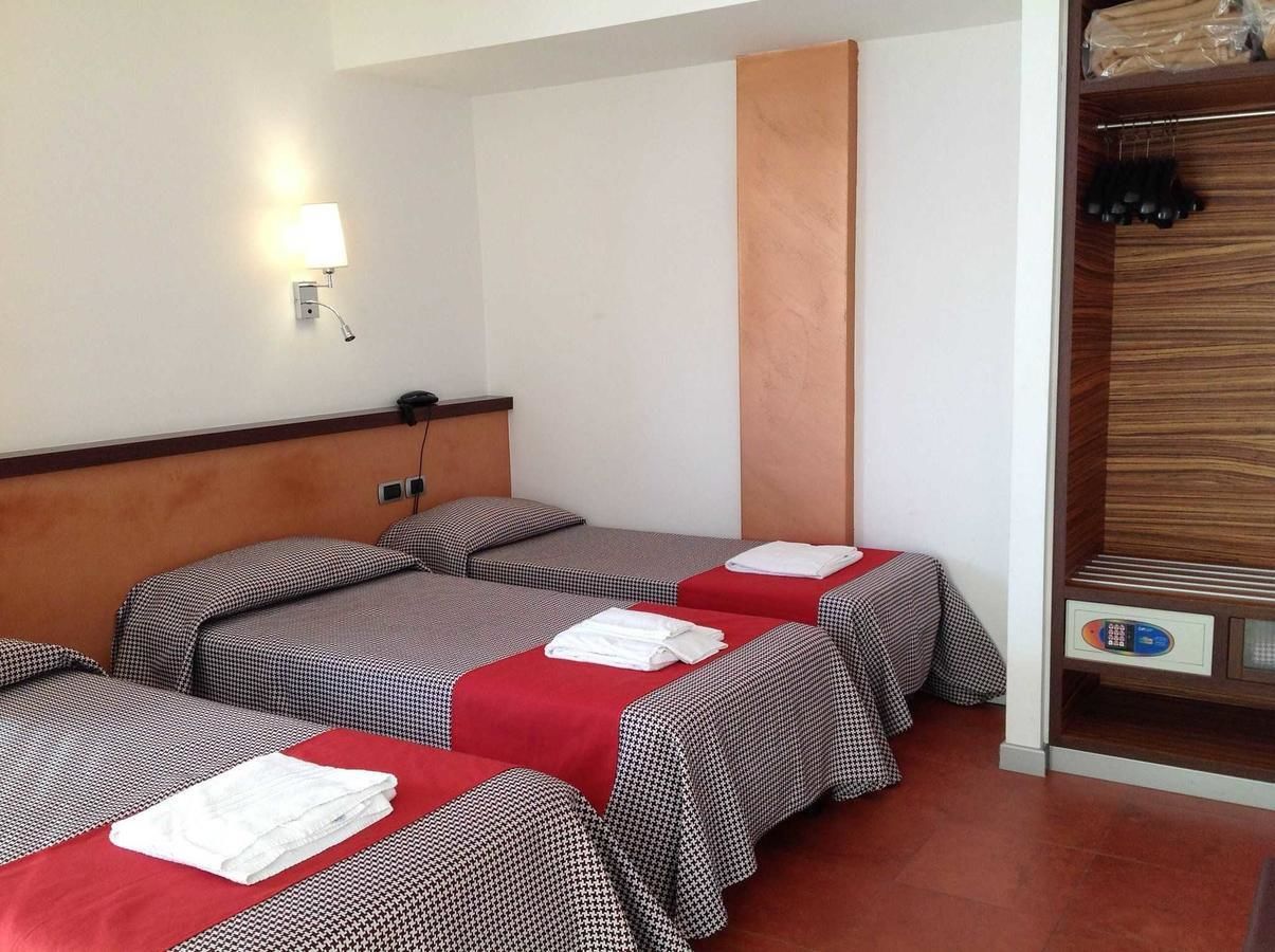 Habitación de hotel con tres camas, cada una con colcha a cuadros rojos y negros y toallas blancas. Cuenta con cabecera de madera, luz empotrada en la pared y un armario de madera con ropa colgada.
