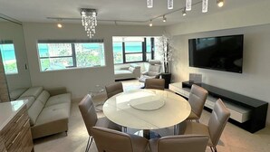 A modern living room with a round dining table, beige sofas, a large TV, and a view of the ocean through the windows. - Beautiful Direct Oceanfront 4 Bedroom with Blue Ocean Views - 702 (Miami Beach)
