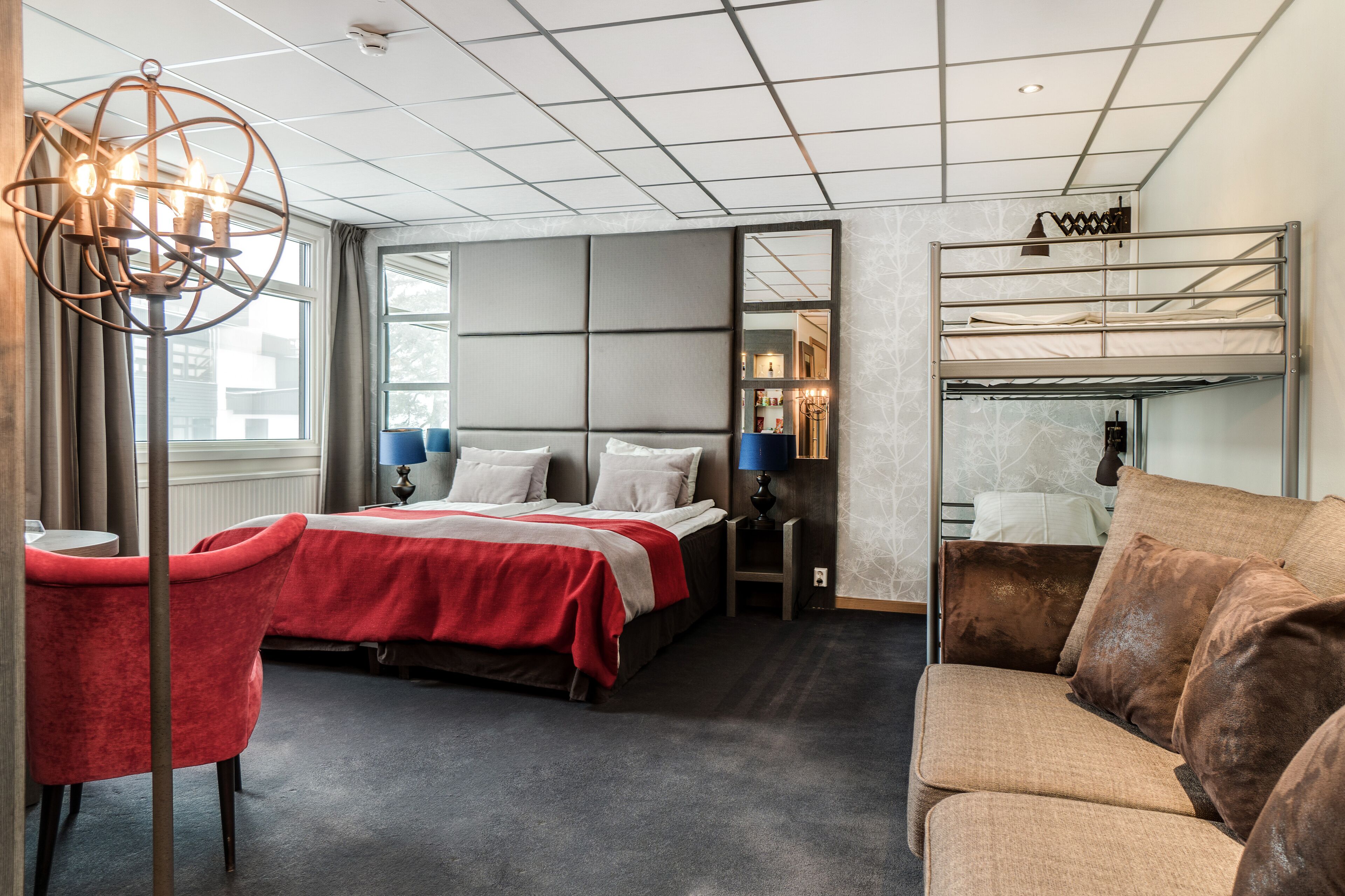 A modern hotel room with a bunk bed, a red chair, a sofa, and a large mirror.