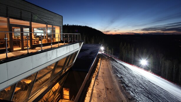 A modern building with large windows, a balcony, and a view of a snowy landscape at dusk. - Hotell Hallstaberget (Solleftea)