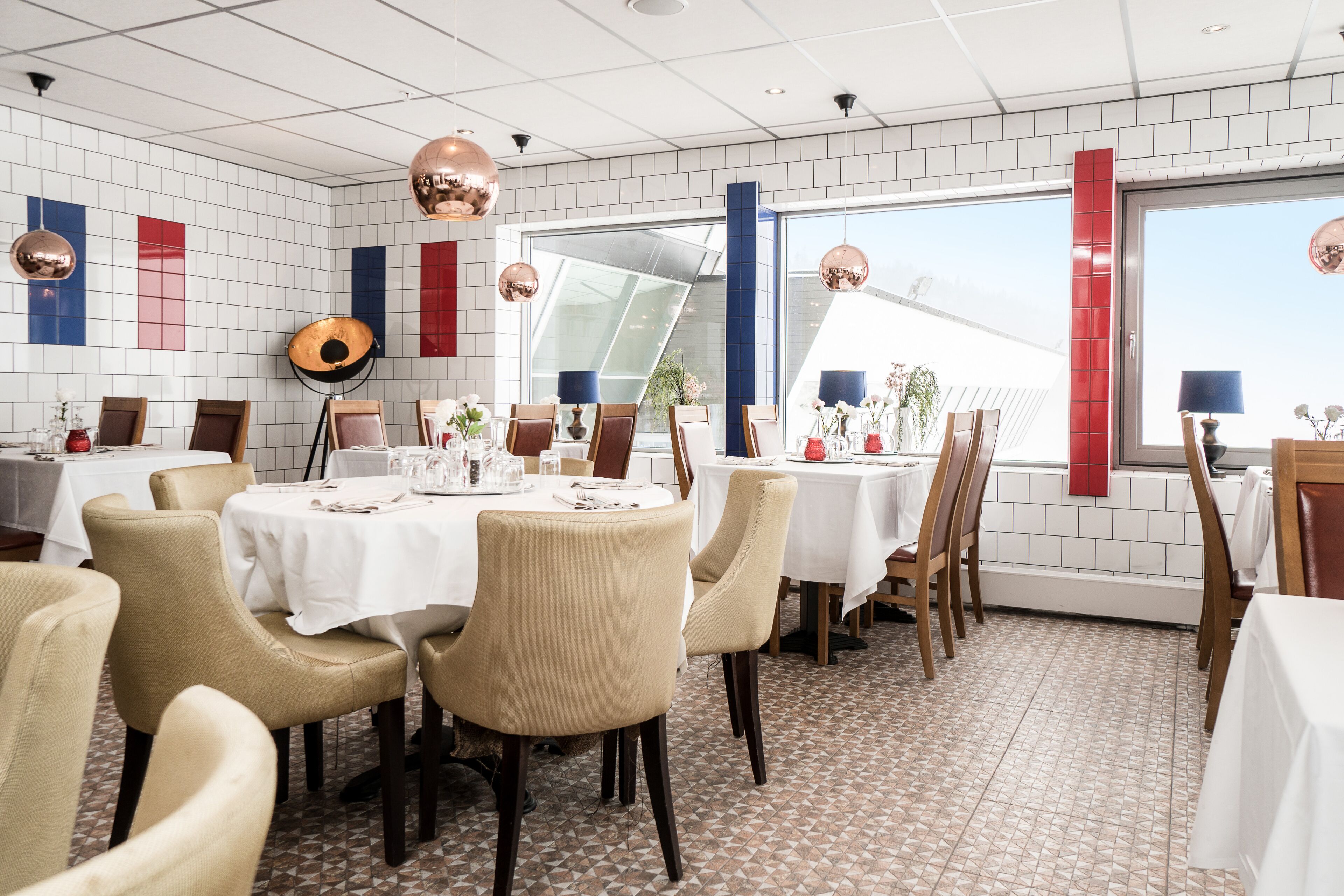 A restaurant with white tiled walls, red and blue accents, and a large window. Tables are set with white tablecloths and chairs.