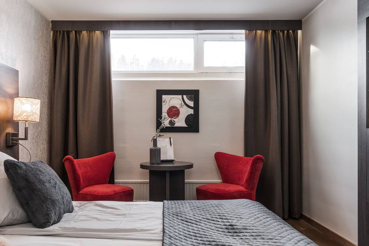 Ein Hotelzimmer mit einem Bett, zwei roten Stühlen, einem kleinen Tisch mit einer Vase und einem gerahmten Bild sowie einem Fenster mit Vorhängen.