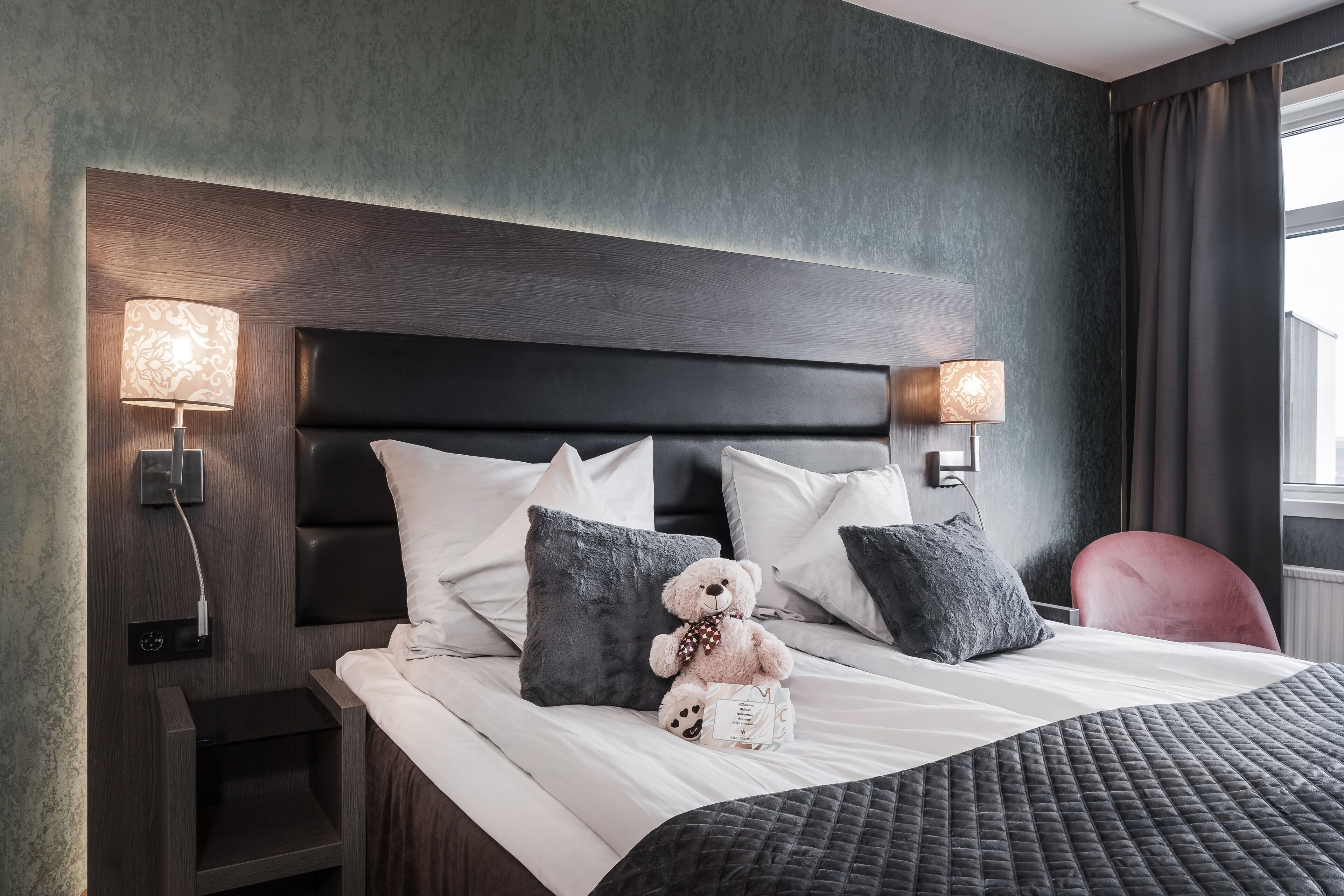A neatly made bed with a dark headboard, two bedside lamps, and a teddy bear on the pillows.