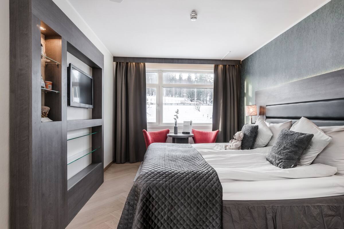 Ein modernes Hotelzimmer mit einem großen Bett, einem an einer dunklen Holzvertäfelung befestigten Fernseher, einem kleinen Tisch mit einer Lampe und Blick auf eine verschneite Landschaft durch ein großes Fenster.