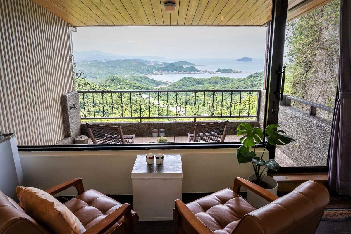 Un balcón con vistas a colinas verdes y una masa de agua. Hay dos sillones de cuero y una mesita con una taza y un platillo.