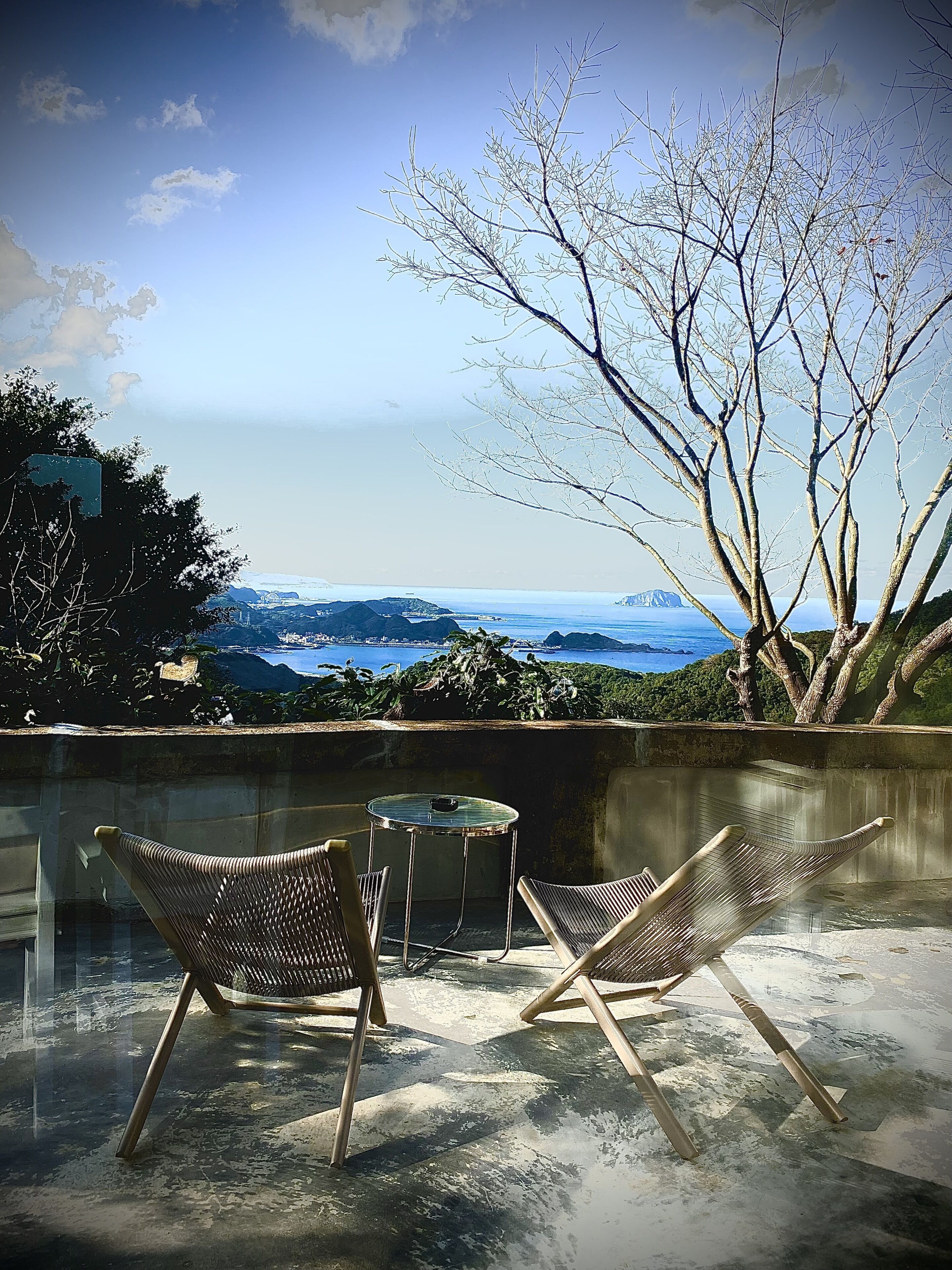 A terrace with two wicker chairs and a small table, overlooking a scenic coastal view with mountains and a tree.