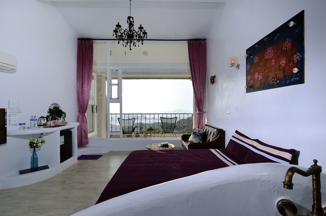 A bedroom with a large bed, a balcony with chairs, a chandelier, and a painting on the wall.