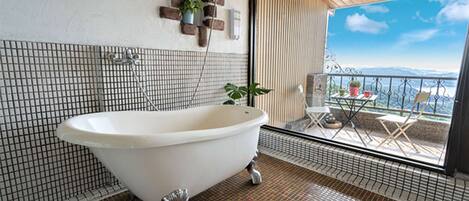 Un baño con una bañera independiente, suelo de baldosas, teléfono en la pared y ventana con vistas a un balcón con mobiliario exterior.
