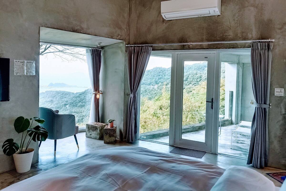 Un dormitorio con una cama grande, una silla, una planta en maceta y vistas a un paisaje montañoso a través de una puerta corredera de vidrio.