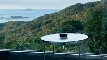 A balcony with a round table and two chairs, overlooking a mountainous landscape with a view of the sea.
