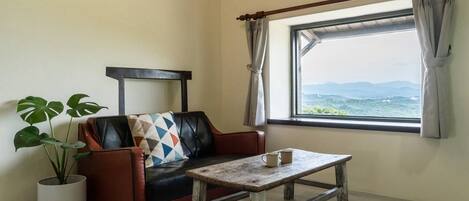 A room with a leather sofa, a wooden table, a potted plant, and a window with a view of mountains.