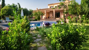 Une maison avec une piscine, une terrasse avec des chaises et une table, et un jardin avec différentes sortes de plantes.