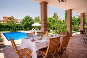 A poolside dining area with a table set for two, surrounded by wooden chairs, a white tablecloth, and a glass of juice. - B&B Villa Marín Toledo (Nambroca)