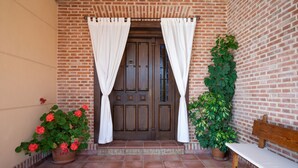 Une entrée d’immeuble en briques avec une porte en bois, des rideaux blancs, un banc et des plantes en pot.