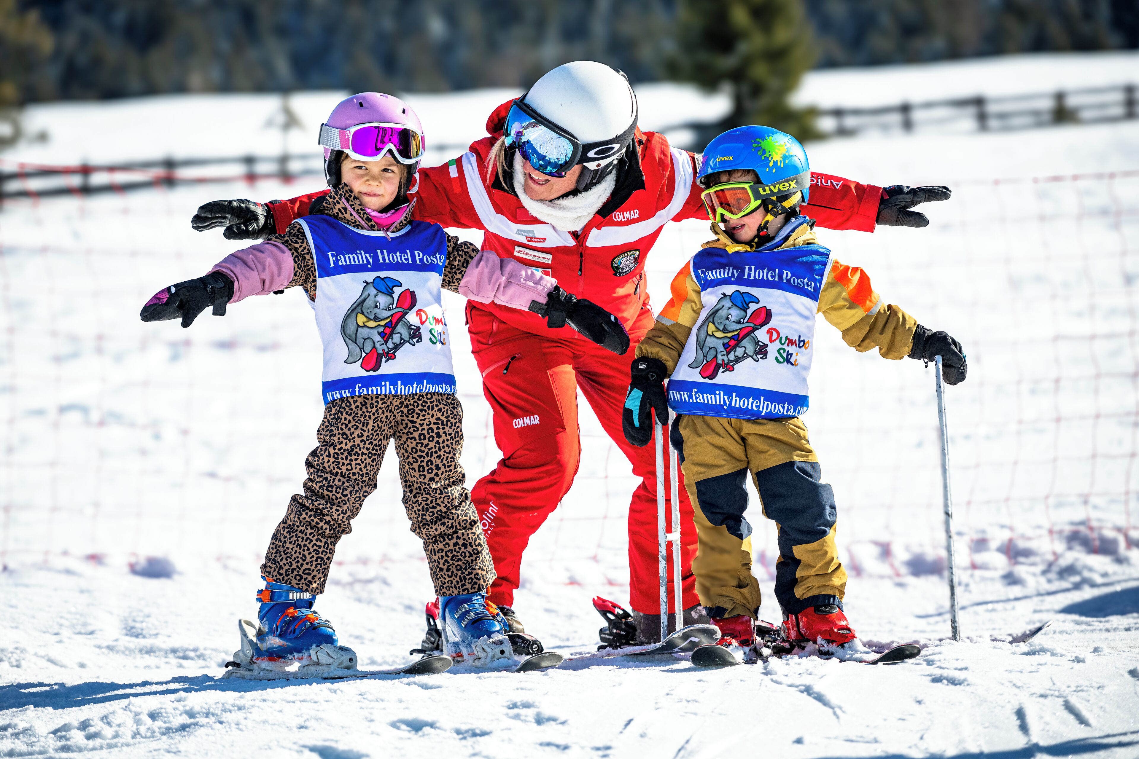 A person in red ski gear with a child in leopard print pants and another child in yellow and blue ski gear.