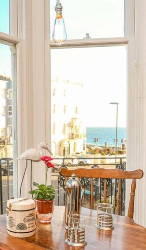 Ein Tisch mit einem Blumentopf, einem Glasbehälter und einer Metallkanne auf einem Balkon mit Blick auf Gebäude und das Meer.
