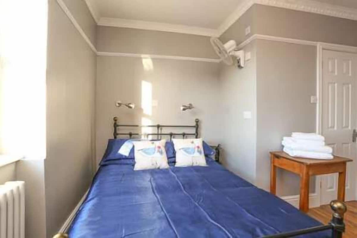 Un lit simple avec un matelas bleu, une tête de lit en métal et deux oreillers. Une petite table en bois avec des serviettes pliées se trouve sur la droite.