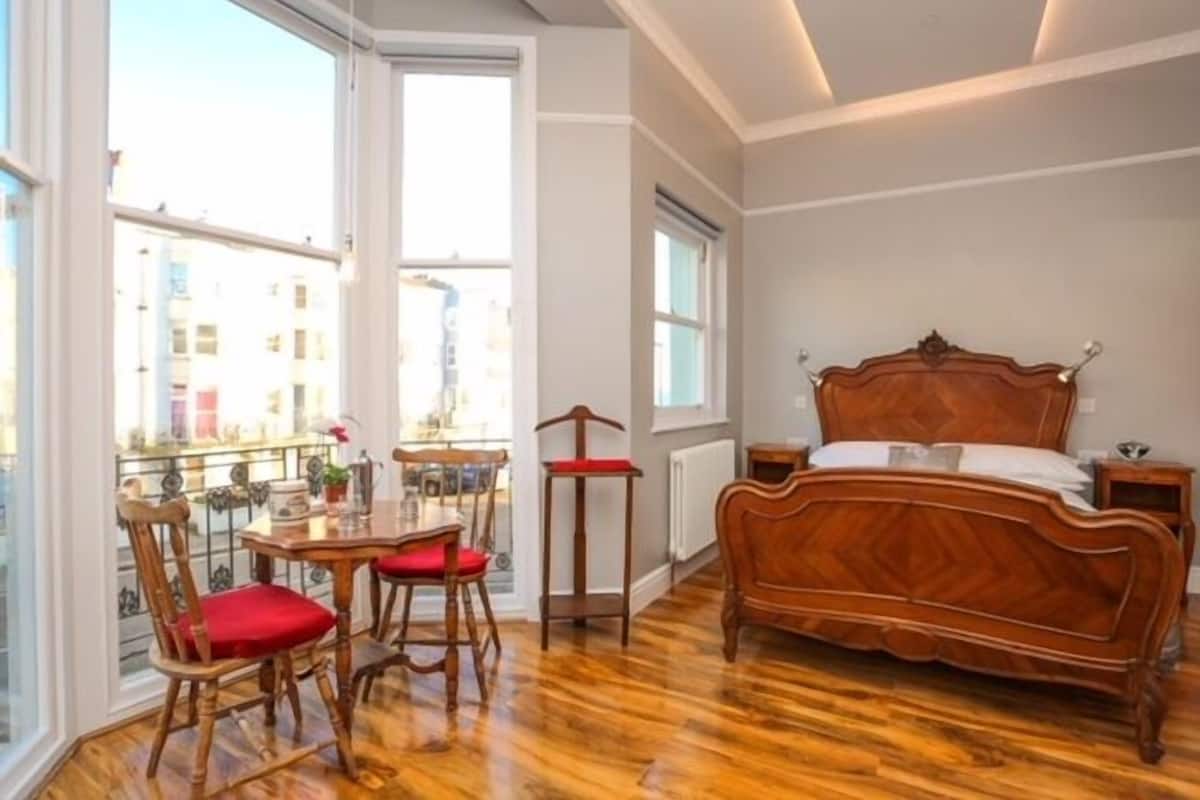 Une chambre à coucher avec un lit en bois, une table à manger avec des chaises et de grandes fenêtres donnant sur des immeubles.