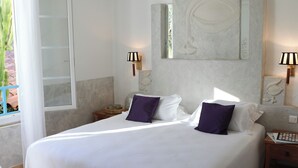 Una cama grande con ropa de cama blanca y almohadas moradas, una mesita de noche con una lámpara y un cuadro abstracto grande en la pared.