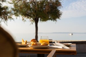 Una mesa con alimentos para el desayuno, incluyendo croissants, un vaso de zumo de naranja y un tazón de sopa, con una vista panorámica de un cuerpo de agua y un árbol.