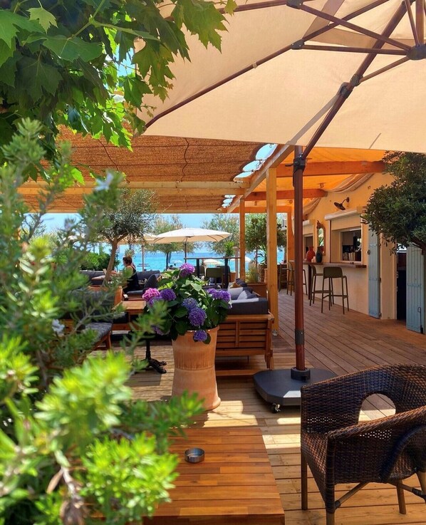 Un espacio al aire libre con mesas y sillas de madera, una maceta grande con flores moradas y vistas al mar.