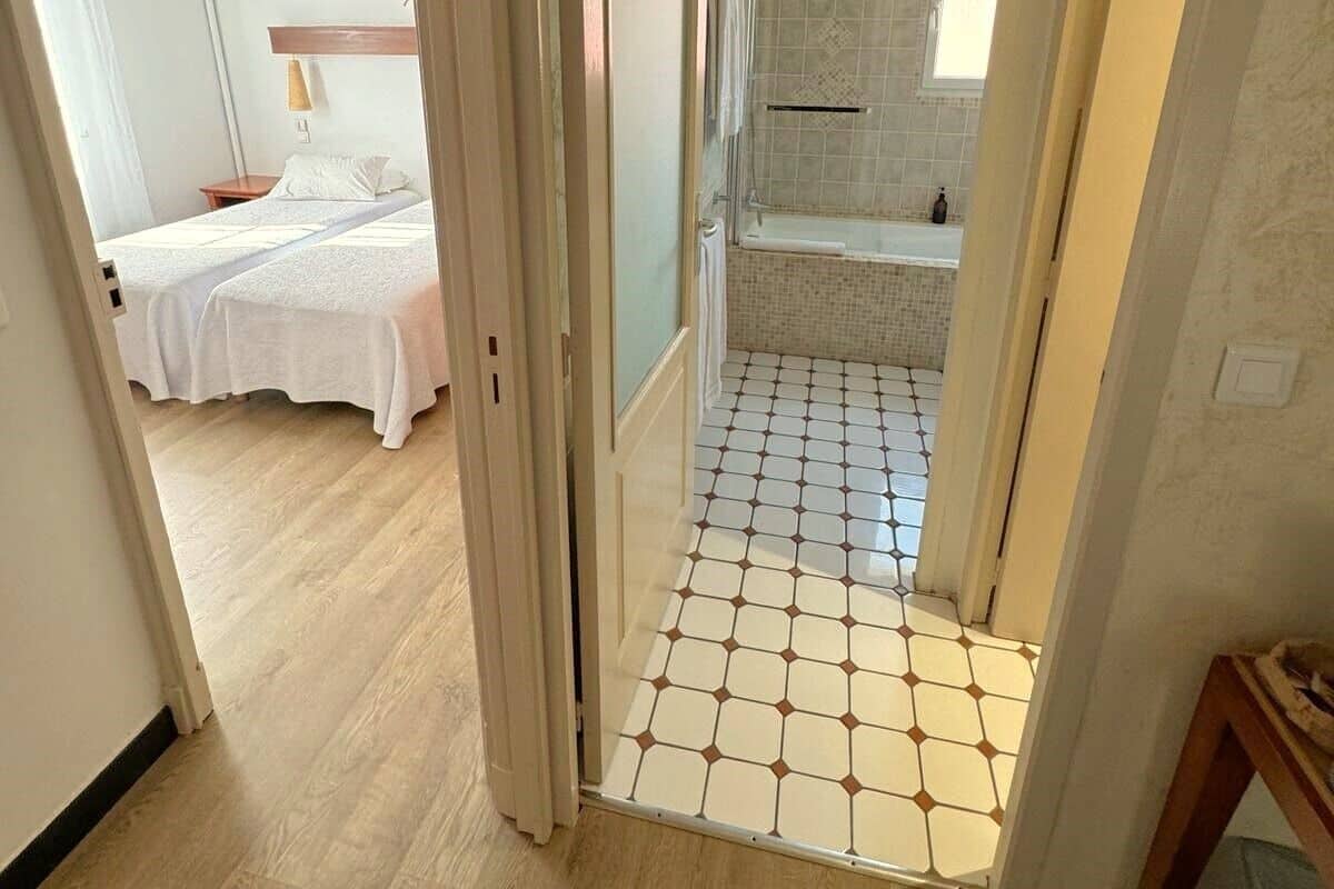 A hotel room with a bed, a bathroom with a shower, and a hallway with tiled flooring.