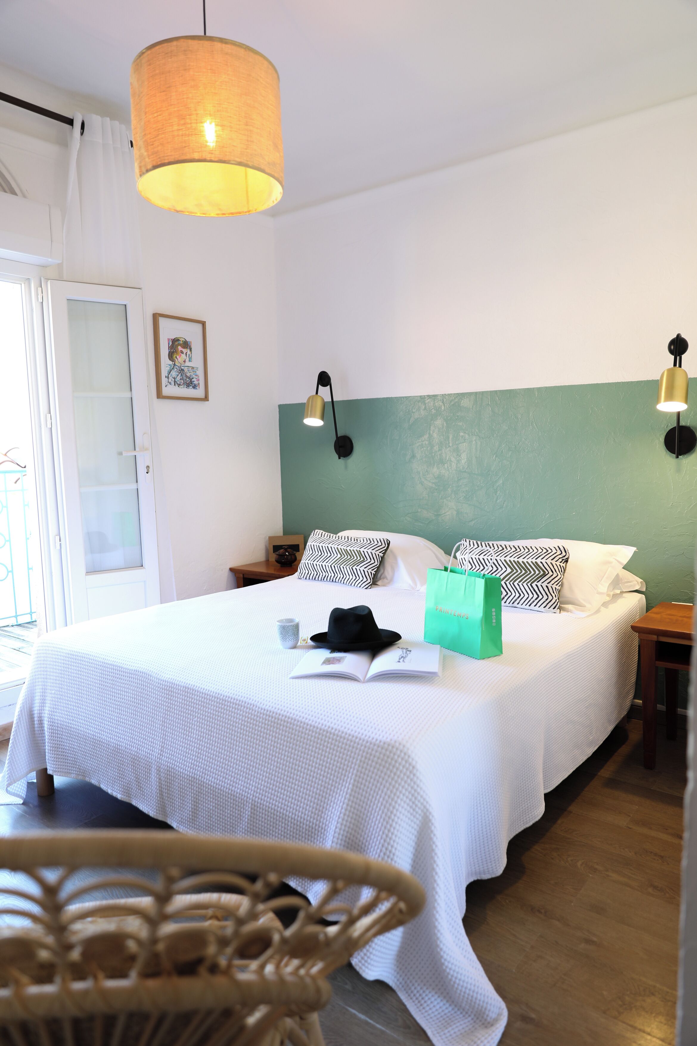 Ein ordentlich bezogenes Bett mit weißer Bettdecke, eine grüne Einkaufstasche und ein schwarzer Hut darauf. Im Vordergrund steht ein Korbstuhl, eine weiße Tür mit Glasfeldern und eine an der Wand befestigte Lampe.