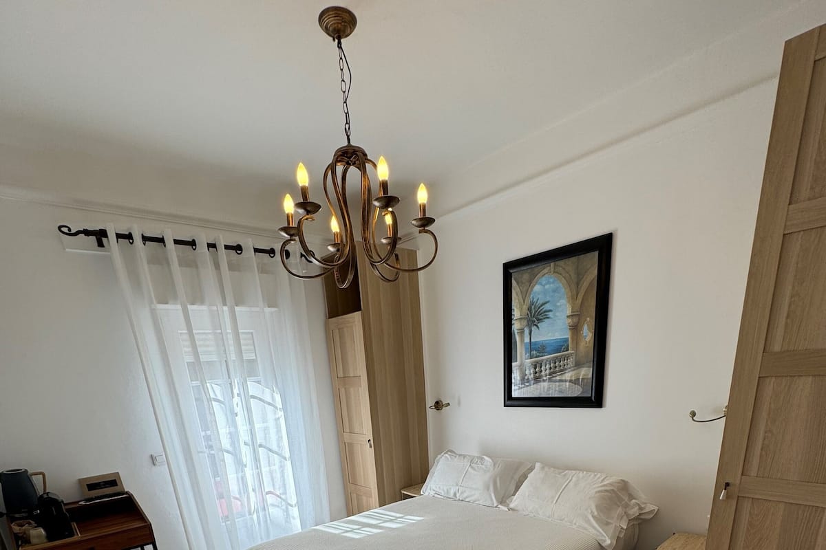 A bedroom with a large bed, a desk, a chair, a wooden wardrobe, and a chandelier.