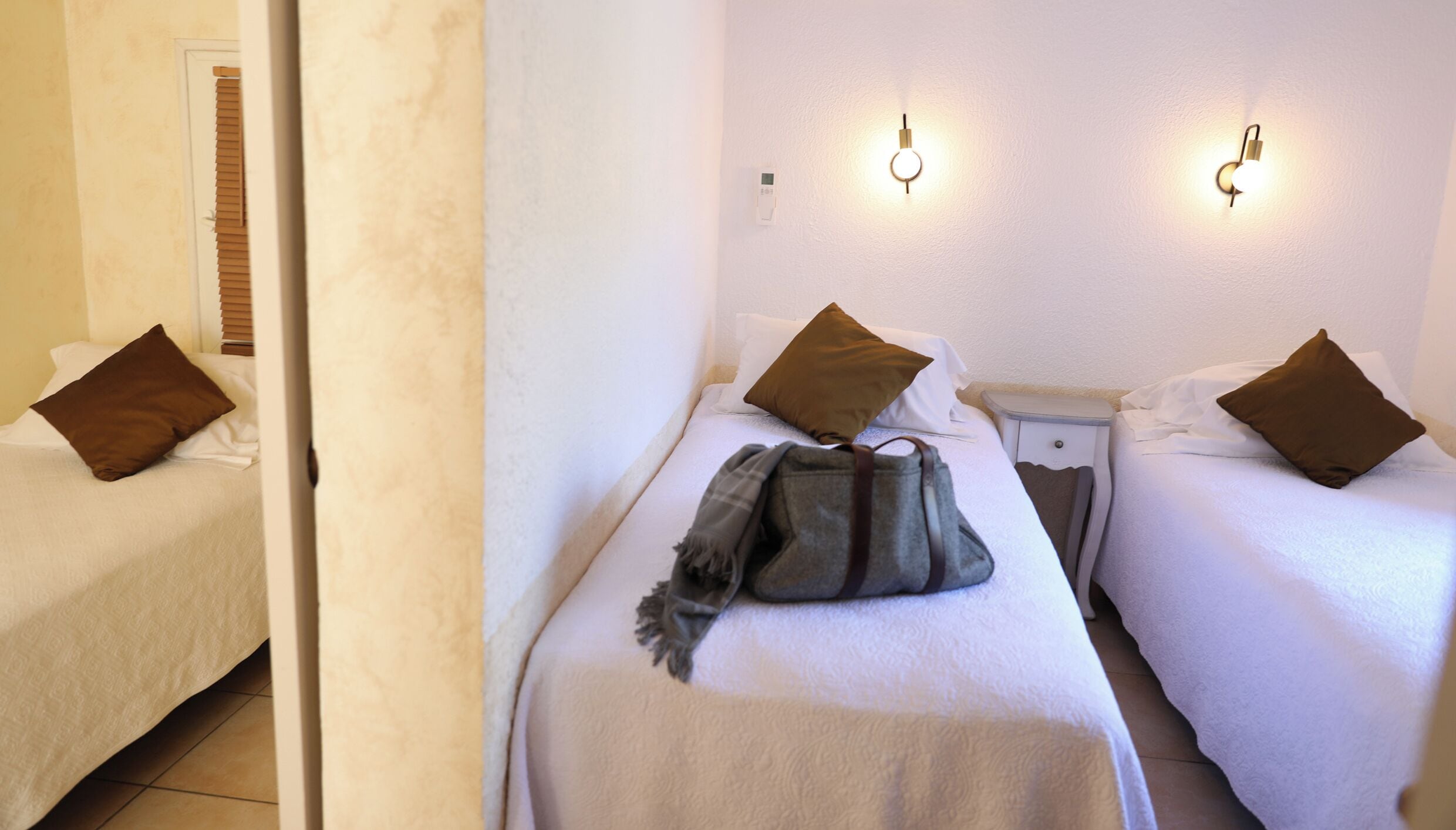 Habitación de hotel con dos camas individuales, cada una con almohada marrón y manta blanca. Se observa una mesita de noche pequeña con una lámpara.