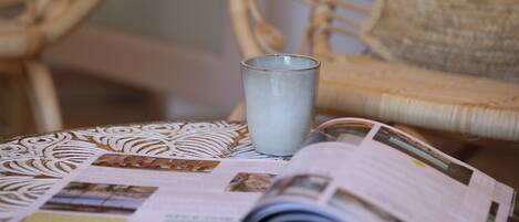 A wicker chair with a cushion, a glass of water, and a magazine on a patterned ottoman.